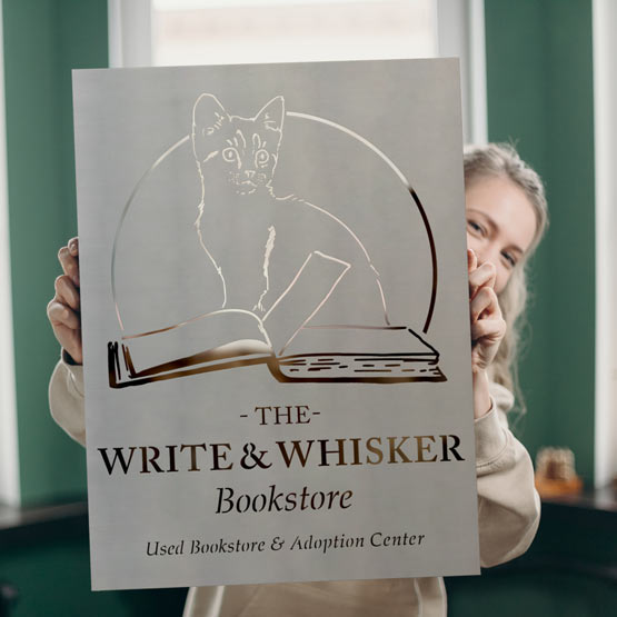 Young Woman Holding Custom Stainless Steel Stencil With Cut Out Of Bookstore Details And Cat Opening Book Logo